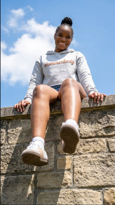 Person wearing a 'Summerbody' hoodie sitting on a stone wall with a blue sky background